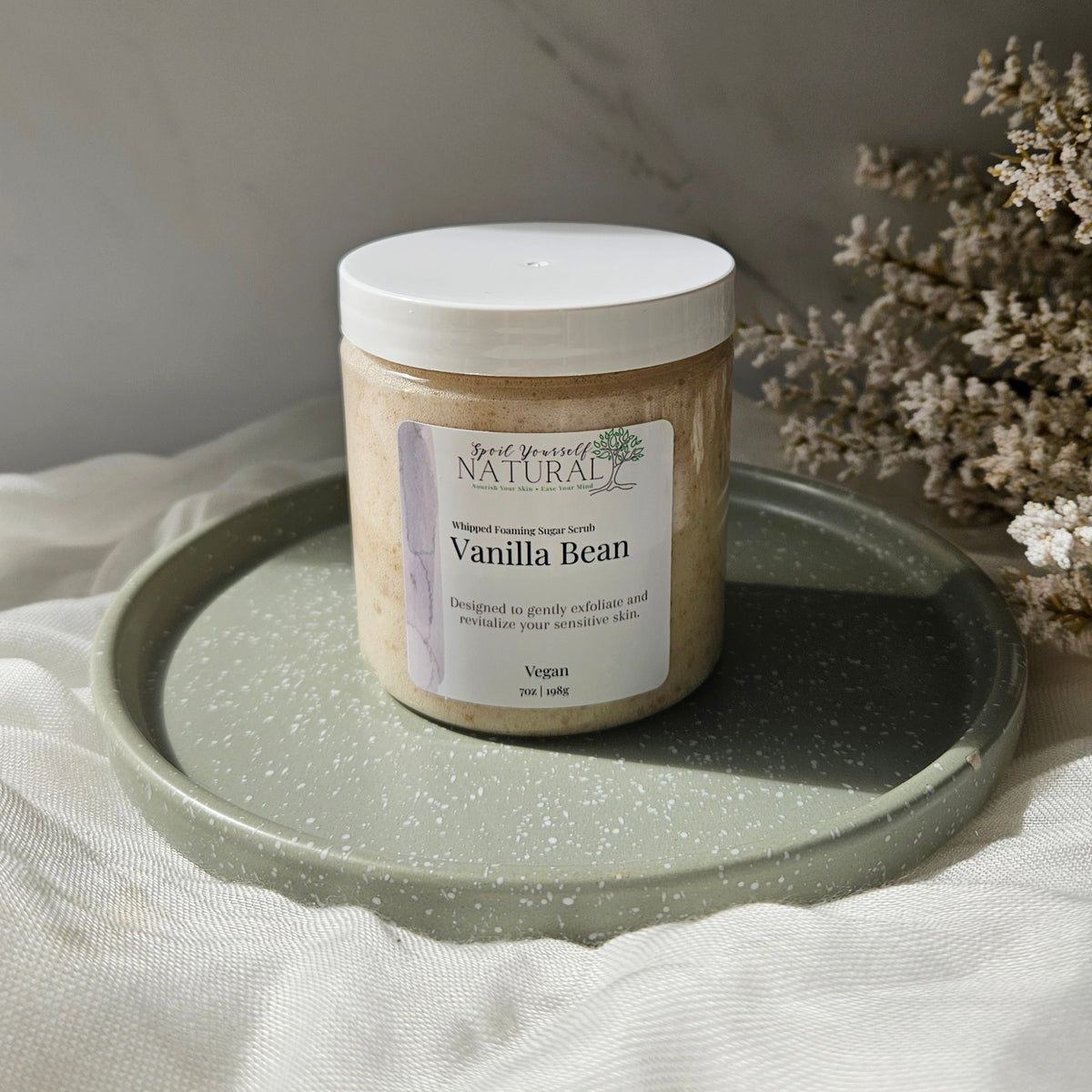 A jar of Vanilla Bean whipped sugar scrub sits on a speckled sage green tray draped with white fabric, with dried flowers in the background.