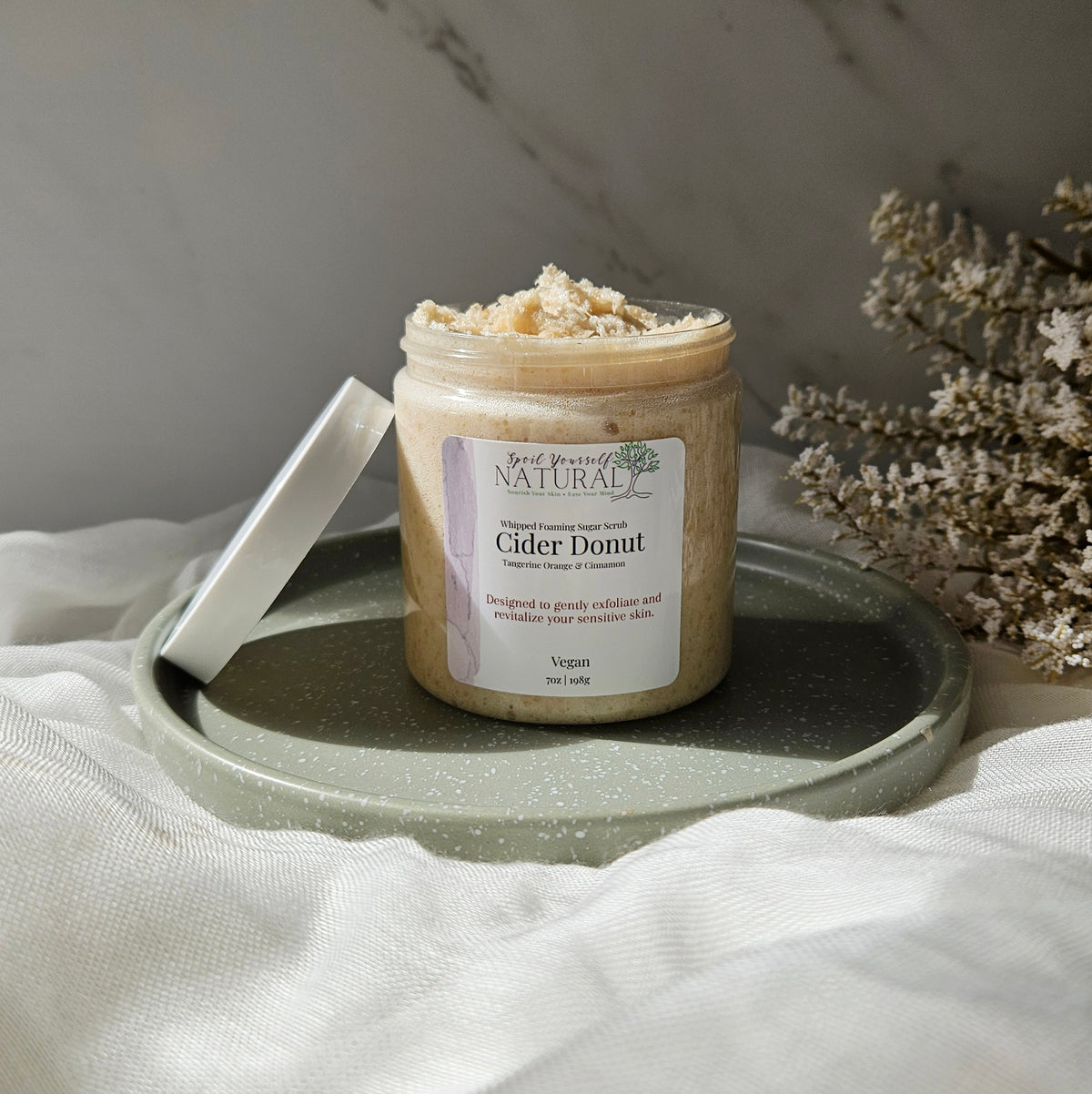 A jar of Cider Donut exfoliating scrub sits on a speckled green tray with its lid leaning beside it. The scene is styled with white fabric and dried flowers against a gray marble background.