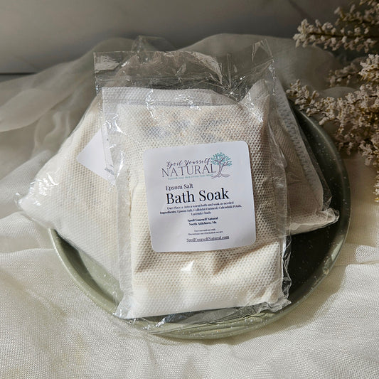 Three packaged bath soak pouches on a dark plate with draped fabric and dried flowers around them. The front pouch has a white label reading 'Hand Made Bath Soak'.