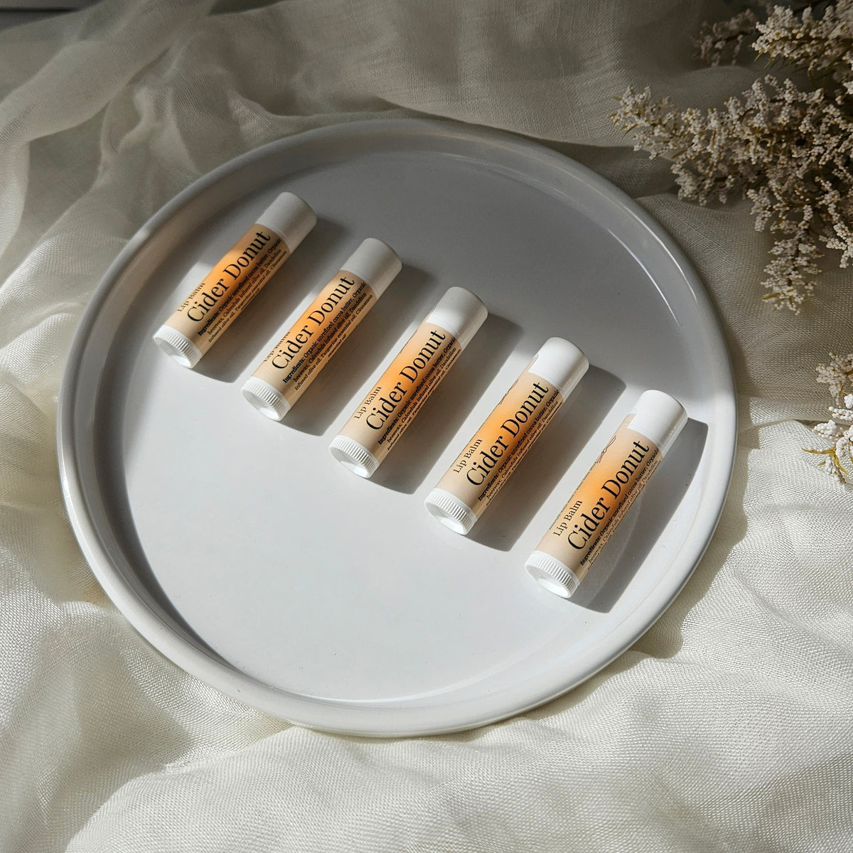 Five lip balm tubes labeled 'Cider Donut' arranged on a round white plate over cream fabric with dried flowers in the corner.