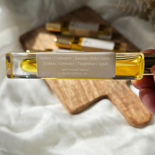 A hand holds a clear glass perfume spray vial containing golden-yellow oil, with a white label listing scents like Amber, Cashmere, Jasmine, Palo Santo, Lemon, Lavender, Tangerine, and Apple. The background is softly blurred with white fabric and a wooden tray holding more similar bottles.