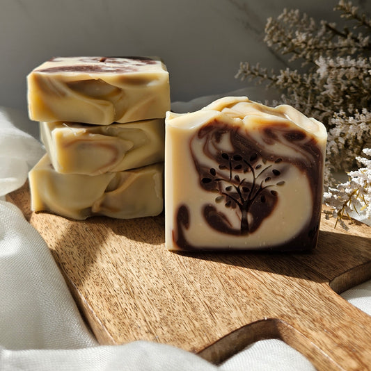 Three stacked bars of handmade soap and one single bar in the foreground on a wooden board, featuring a cream and dark brown marbled swirl pattern with botanical leaf details, styled with white fabric and dried flowers.