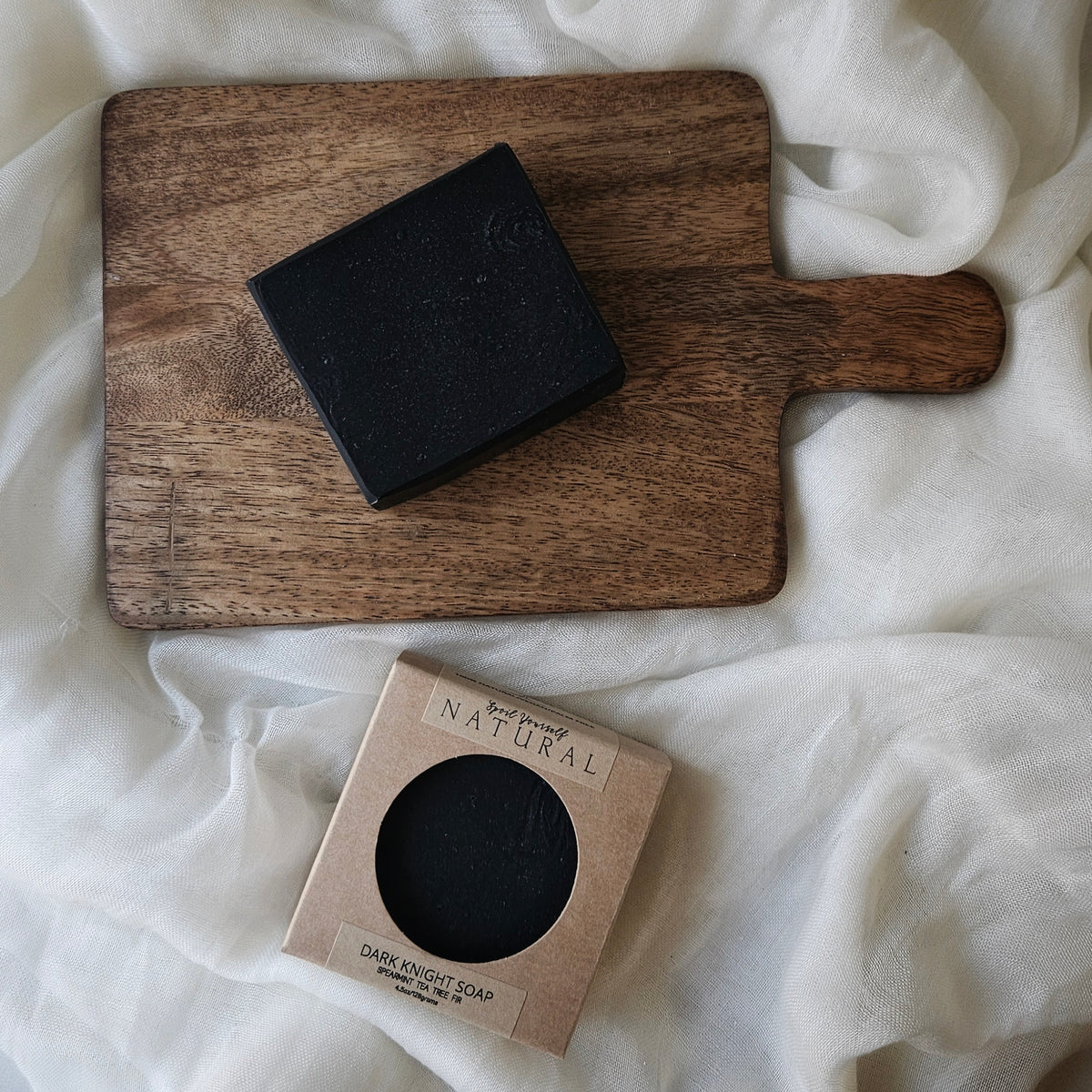 A black bar of soap resting on a wooden board with a box packaging beside it, all placed on a white cloth surface.