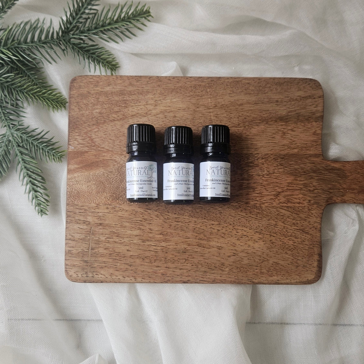 Three small black bottles labeled 'Natura' on a wooden cutting board with greenery on a light fabric background.