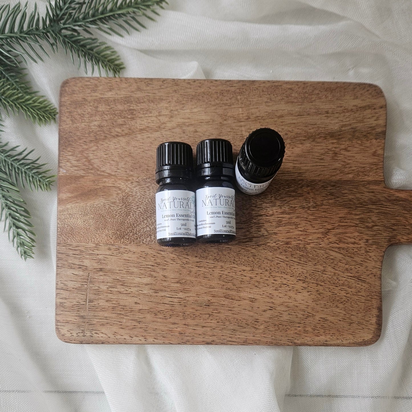 Three small bottles of essential oils on a wooden cutting board with greenery on a white fabric background