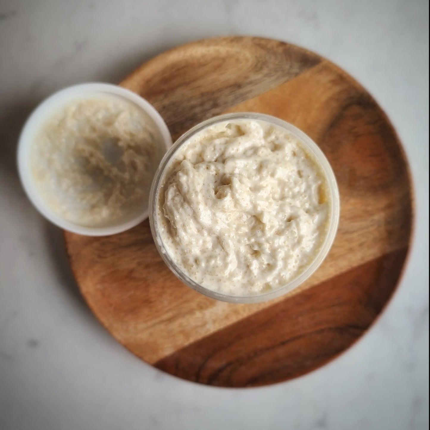 A bowl of sugar scrub is placed on a wooden plate with a spoon beside it, indicating a creamy texture.