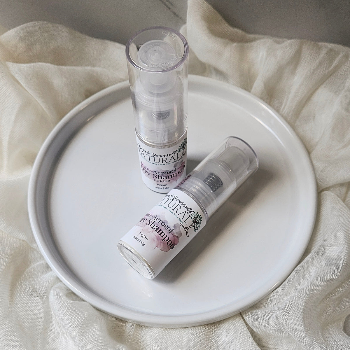 A close up of a bottle of non-aerosol dry shampoo placed on a white surface, with a cap beside it, against a marble background.