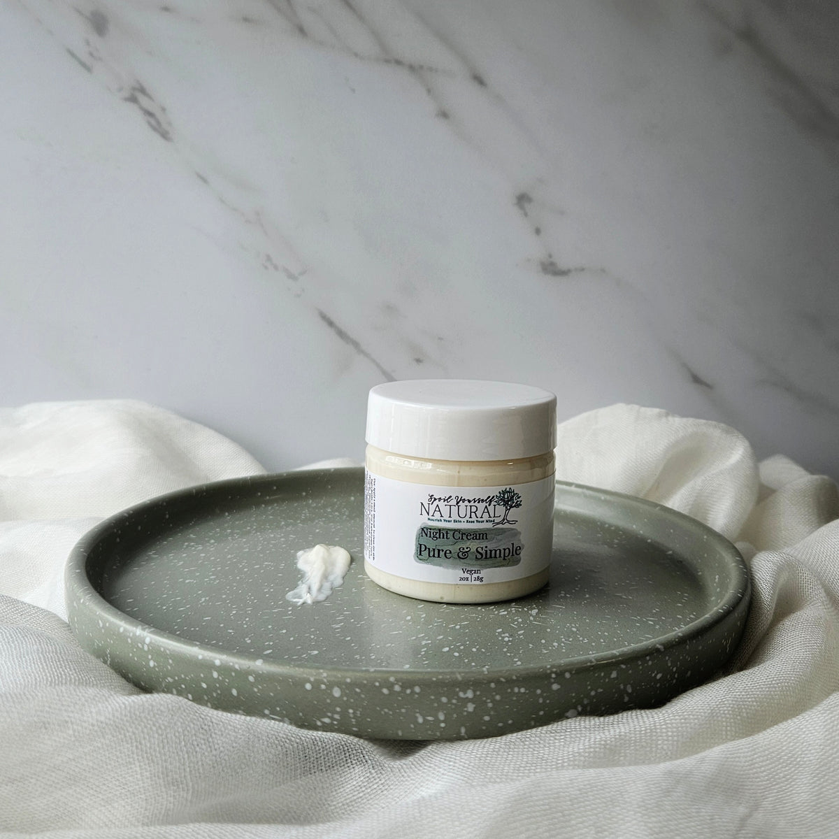 A jar of facial night cream placed on a ceramic plate with a small amount of cream spread out in front of it, all set against a marble background with a neutral color scheme.