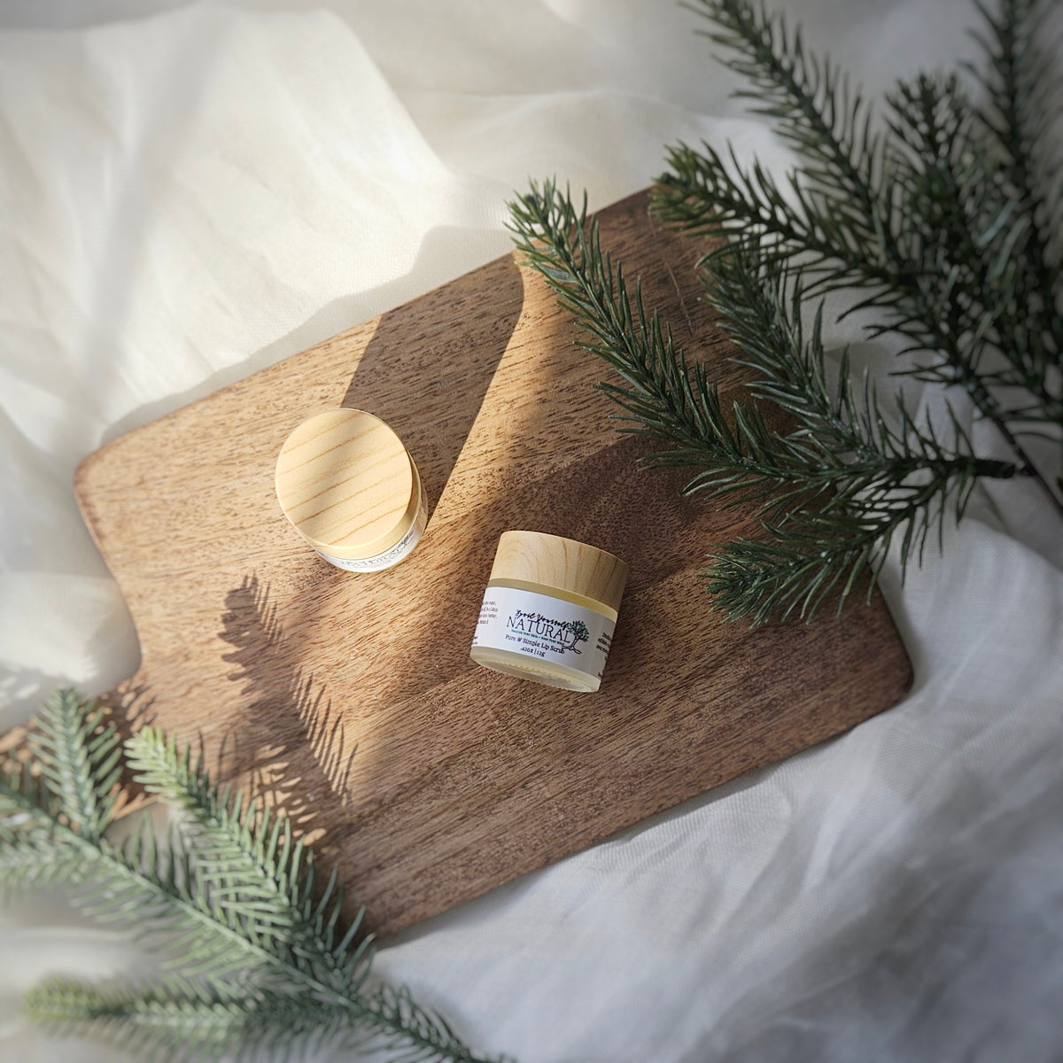 A small jar of unscented lip scrub is placed on a wooden surface with a few drops of oil, beside greenery that suggests a natural or outdoor setting.