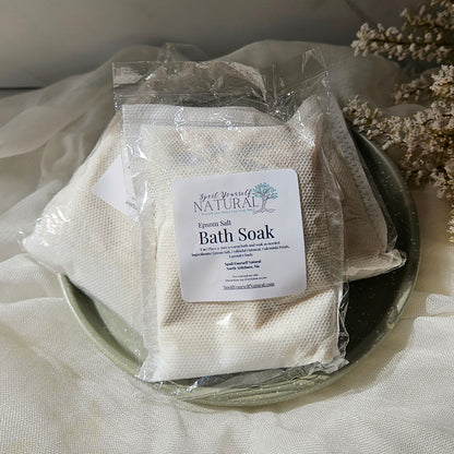 Three packaged bath soak pouches on a dark plate with draped fabric and dried flowers around them. The front pouch has a white label reading 'Hand Made Bath Soak'.