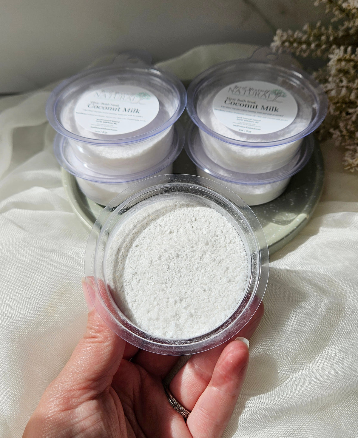 A hand holds a clear round container filled with white granular scrub in front of three more stacked containers labeled Coconut Milk on a draped white fabric surface.