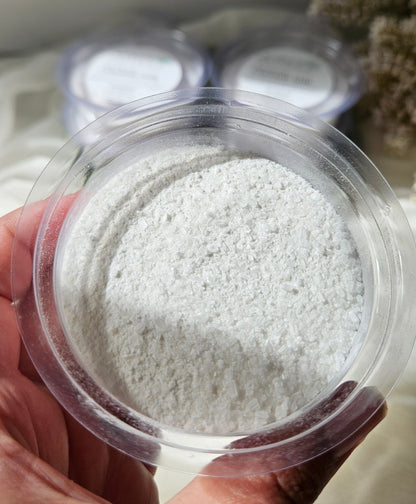 A hand holding a clear round plastic jar filled with white powder, with two more similar jars blurred in the background.