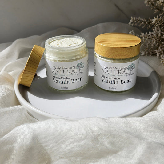 Two jars of Whipped Tallow Vanilla Bean shown on a white ceramic tray with draped cream fabric. One jar is open, showing the white whipped tallow inside, and the other has a bamboo lid resting beside it.
