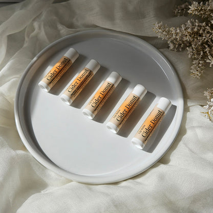 Five lip balm tubes labeled 'Cider Donut' arranged on a round white plate over cream fabric with dried flowers in the corner.