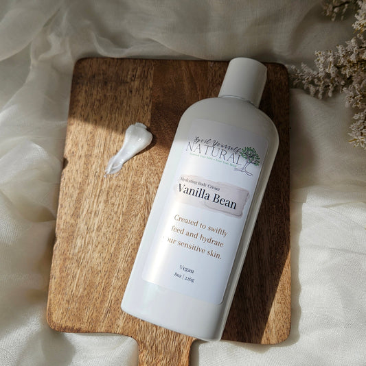 A bottle of Vanilla Bean moisturizer is displayed on a wooden board with a dollop of white cream next to it, set against a draped white fabric background.