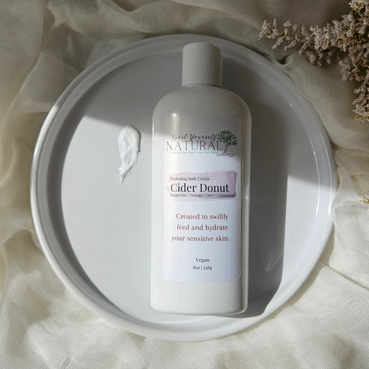 A bottle of  Cider Donut Body Cream sits on a white ceramic plate, surrounded by draped cream fabric and dried flowers.