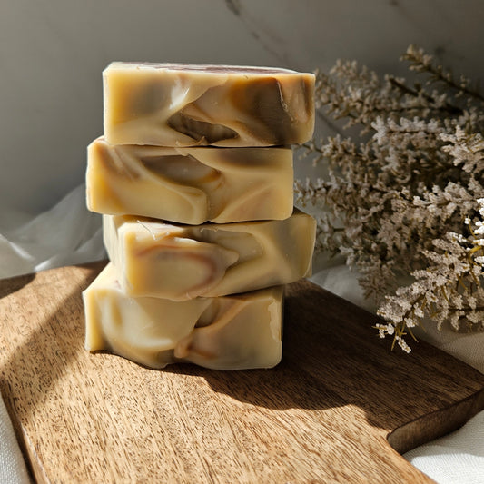 Four stacked rectangular bars of handmade soap with a marbled cream and brown swirl pattern, displayed on a wooden cutting board with dried white flowers in the background.