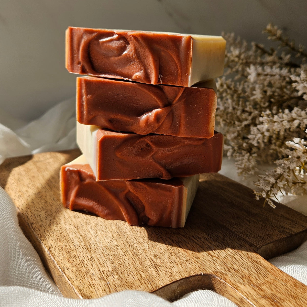 Four stacked rectangular bars of handmade soap with a swirled terracotta-brown and cream pattern, displayed on a wooden cutting board with dried flowers in the background.