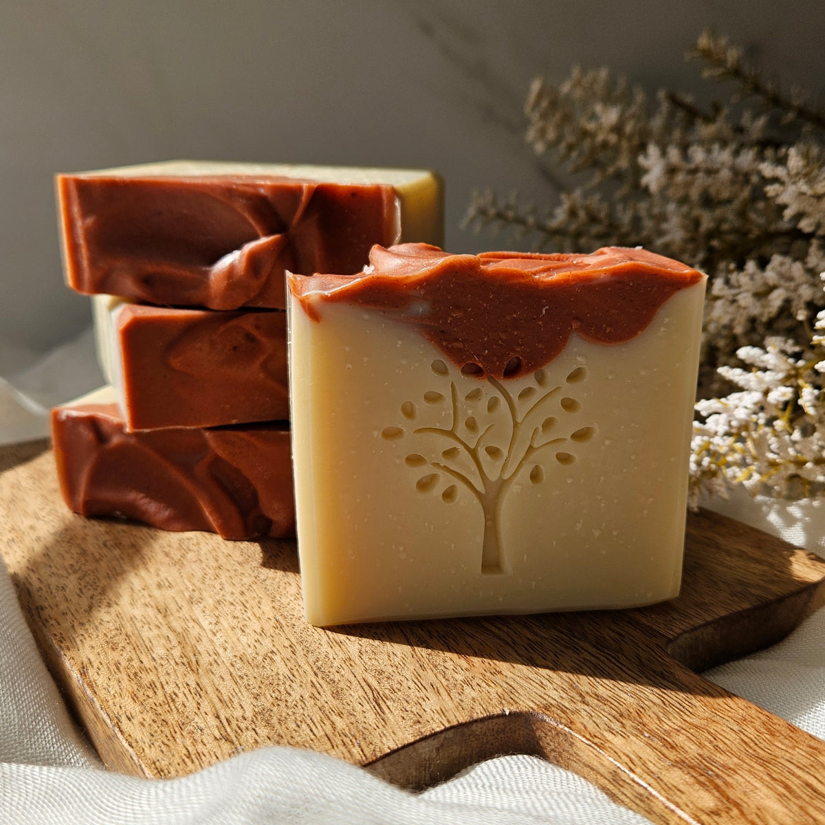 Three handmade rectangular bars of soap displayed on a wooden board, with one bar in front showing an embossed tree design and a swirled reddish-brown top layer, set against a soft neutral background with dried flowers.