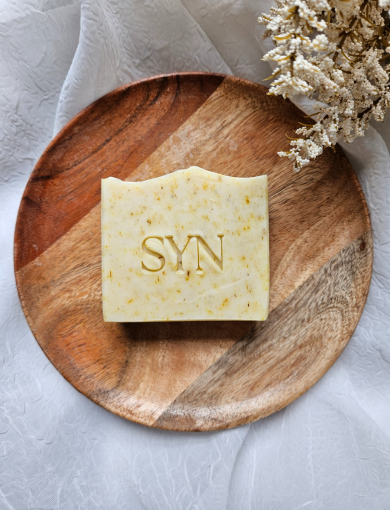 A bar of Calendula Aloe & Oats Soap placed on a wooden plate with a light-colored powder scattered around, possibly kaolin clay.