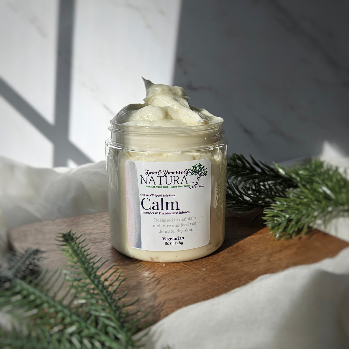 A jar of 'Calm' whipped body butter by Spoil Yourself Natural, with a marble background and pine branches in the foreground.