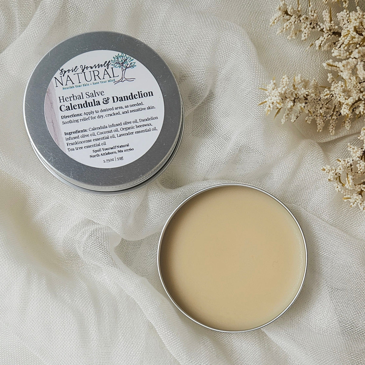 A metal tin with lid open, displaying a beige herbal salve labeled 'Good Friend Natural - Herbal Salve Calendula & Dandelion'. The tin is placed on a white fabric alongside dried plants.