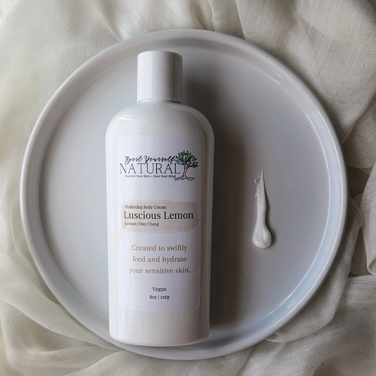 Body cream bottle labeled 'Luscious Lemon' on a white plate with a beige fabric background