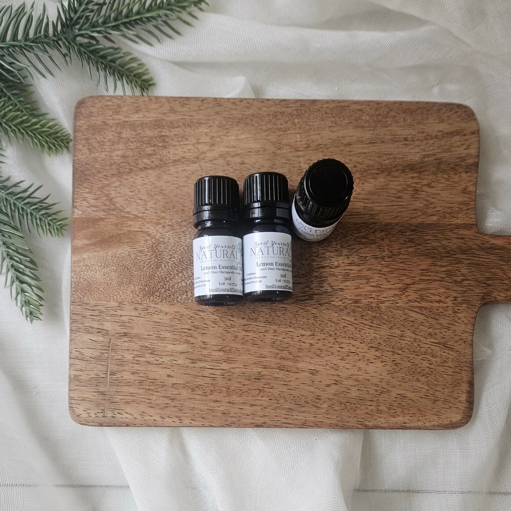 Three small bottles of essential oils on a wooden cutting board with greenery on a white fabric background