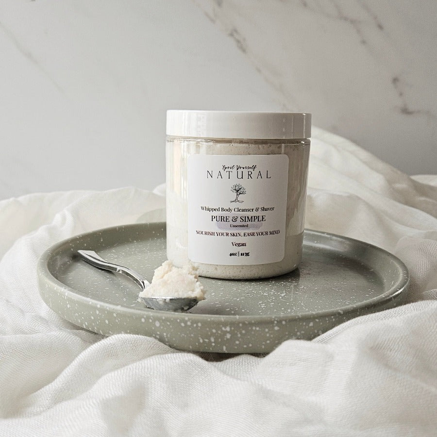 A jar of Pure & Simple Whipped Body Cleanser & Shaver on a grey plate with a spoonful of the product beside it, all placed on a white towel with a marble background.