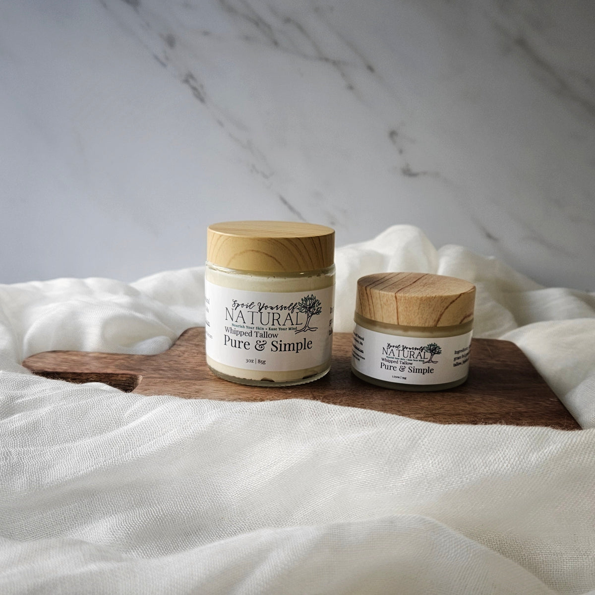 Two containers of whipped tallow balm, one larger and one smaller, with labels that read 'Nature's Pure Simple, Fresh & Simple'. They are placed on a white fabric surface with a marble background.