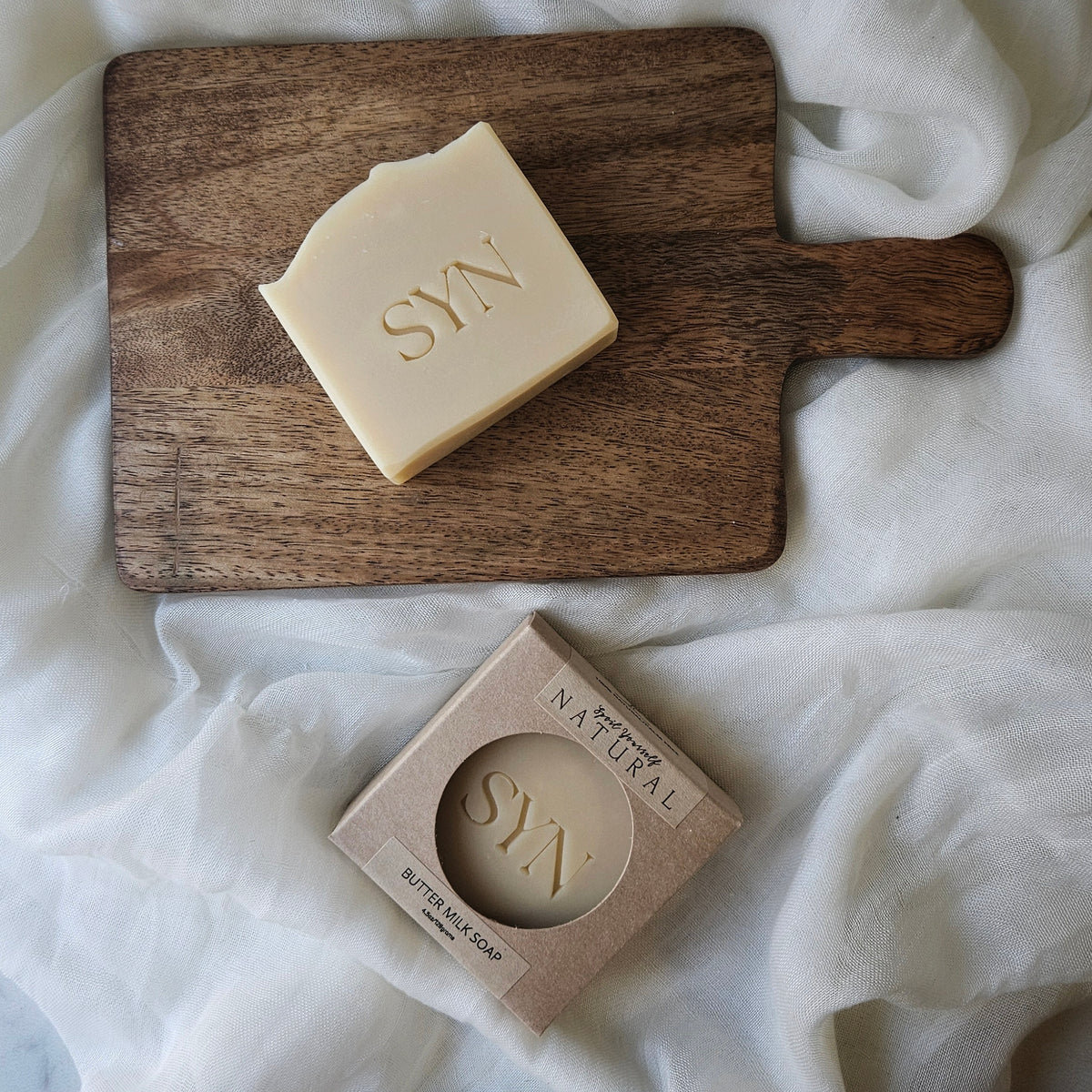 A bar of Butter Milk Soap resting on a wooden board with a light-colored cloth beneath it. A box with the soap visible is also shown, bearing the brand name 'SYN'.