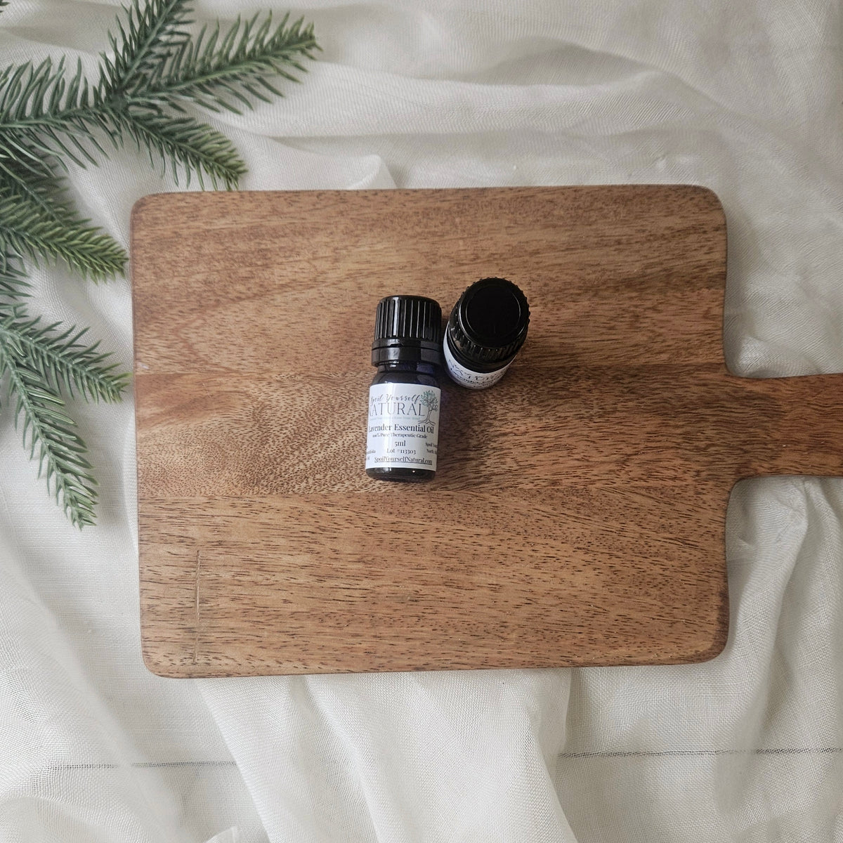 Two essential oil bottles on a wooden cutting board with greenery on a white fabric background