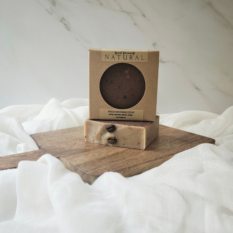 A bar of soap with a brownish color and exfoliating particles, placed on a wooden surface with a box on top that has a transparent window showing the product.
