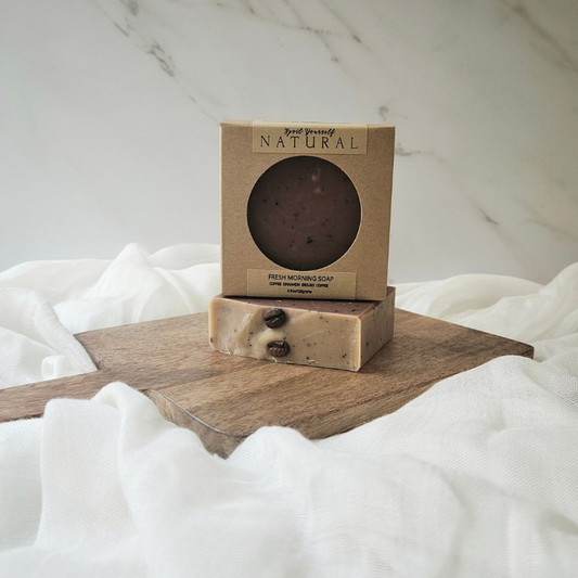 A bar of soap with a brownish color and exfoliating particles, placed on a wooden surface with a box on top that has a transparent window showing the product.