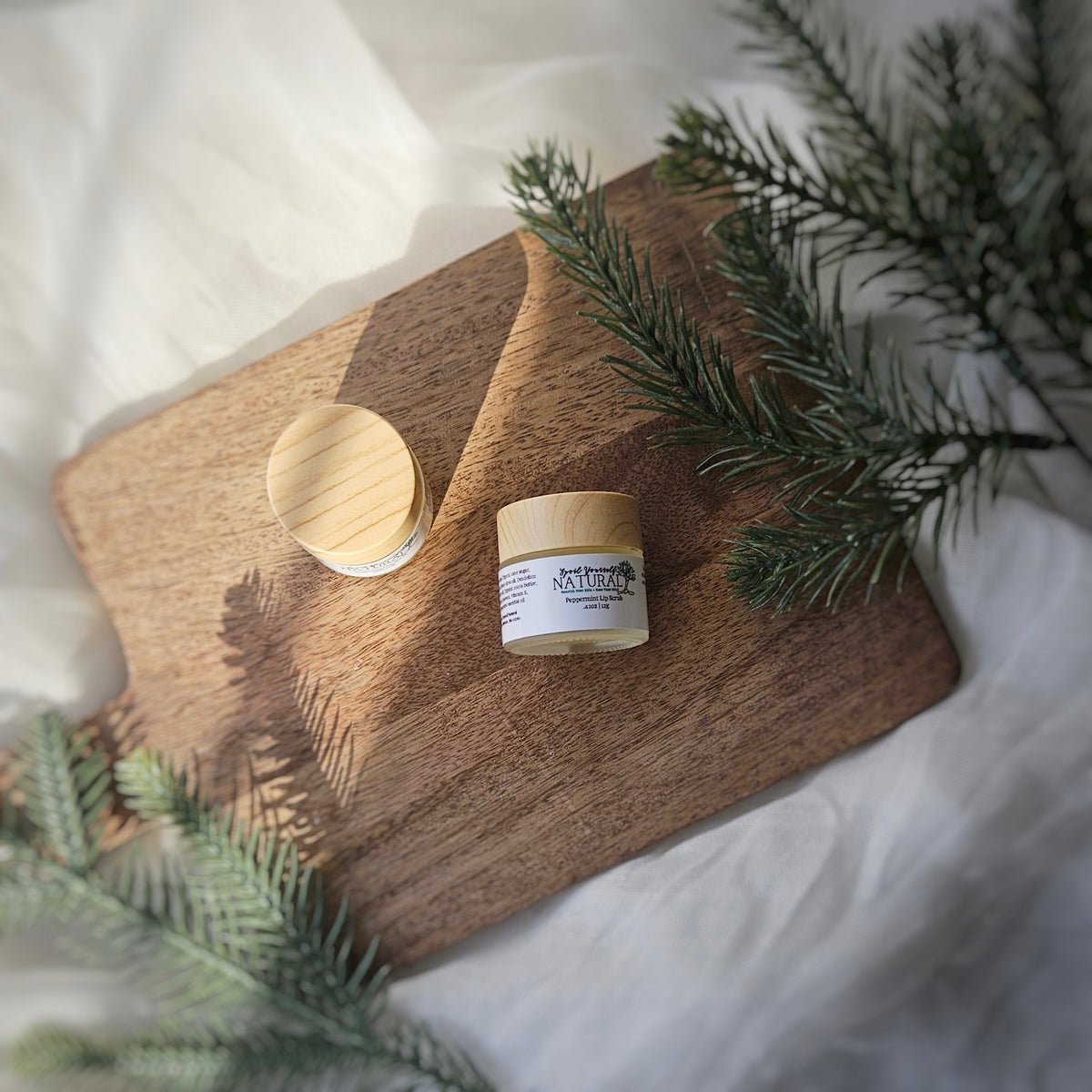 A small jar of peppermint lip scrub is placed on a wooden surface with a few drops of oil, beside greenery that suggests a natural or outdoor setting.