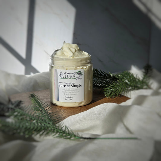 A jar of 'Pure & Simple Whipped Body Butter' by Spoil Yourself Natural, placed on a wooden surface with a shadow falling over it, surrounded by decorative greenery.