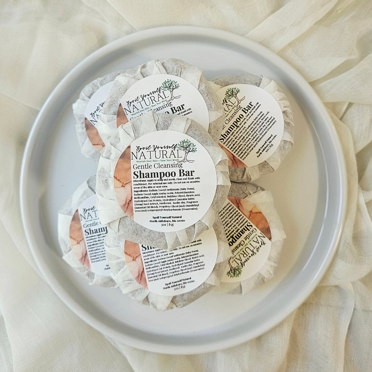Multiple shampoo bars wrapped in individual paper wrappers, placed on a white plate with a soft, textured background.