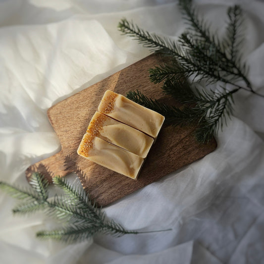 A bar of Spiked Orange soap on a wooden cutting board, with a piece partially sliced to show the creamy interior, placed on a soft, white fabric background with green pine needles around.