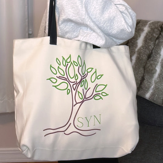 A beige custom tote bag with a green tree and the letters 'SYN' printed on the front, resting on a couch with a grey blanket in the background.