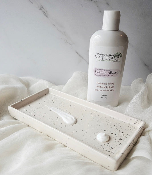 A bottle of SYNfully Aligned Cream with its cap removed, placed next to a small amount of cream on a white ceramic dish, all presented on a soft, textured white surface.