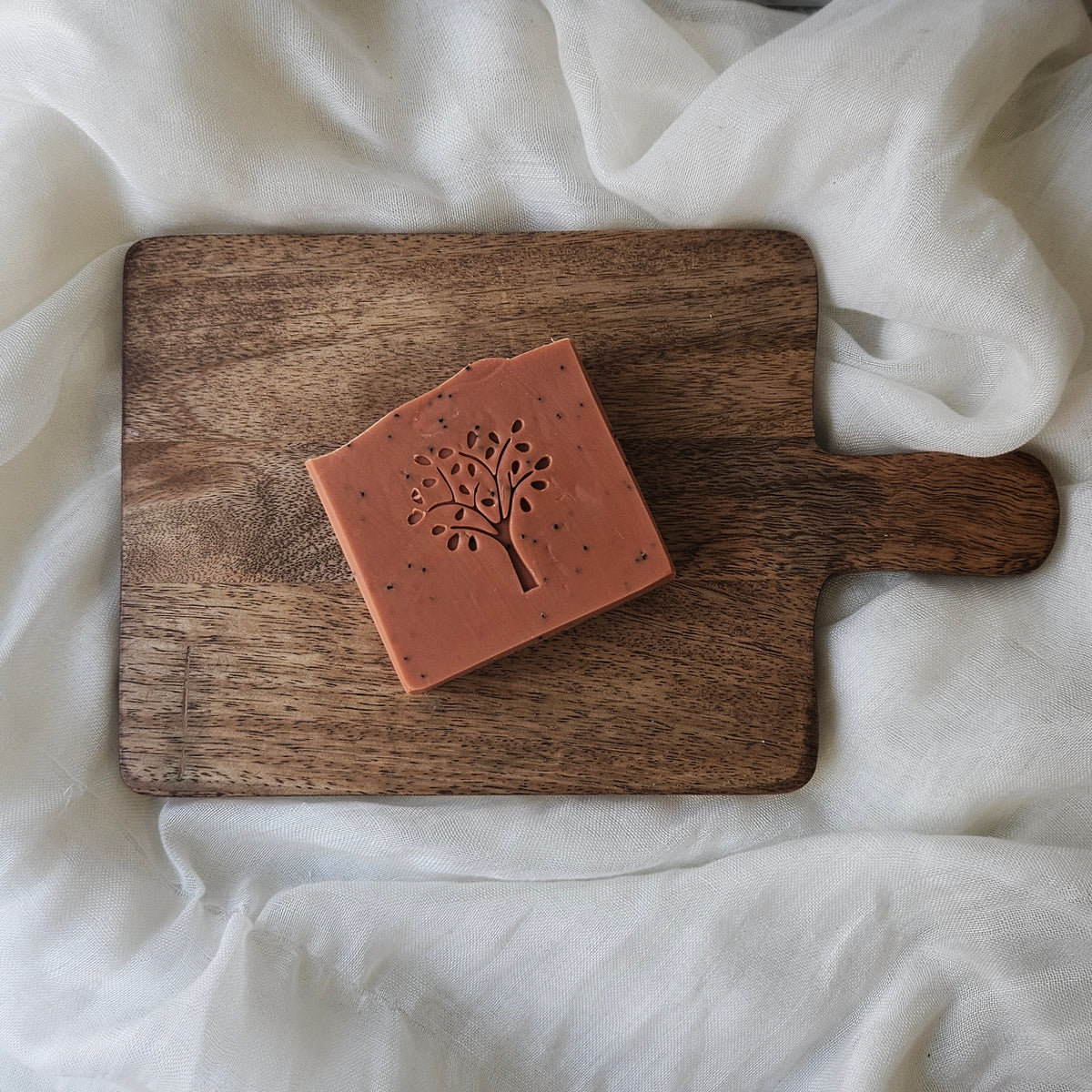 A bar of soap with a tree design on top, placed on a wooden board with a white cloth in the background.