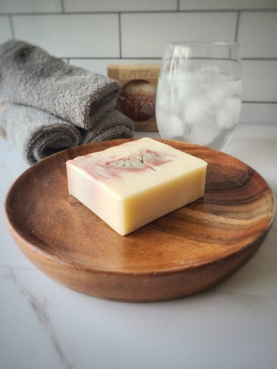 a bar of fresh and clean soap sitting on a wooden dish with facial clothes and a glass of water in the background