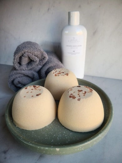 Three lemon-colored bath bombs in a green dish with a grey towel and a white bottle in the background, suggesting a spa-like setting.
